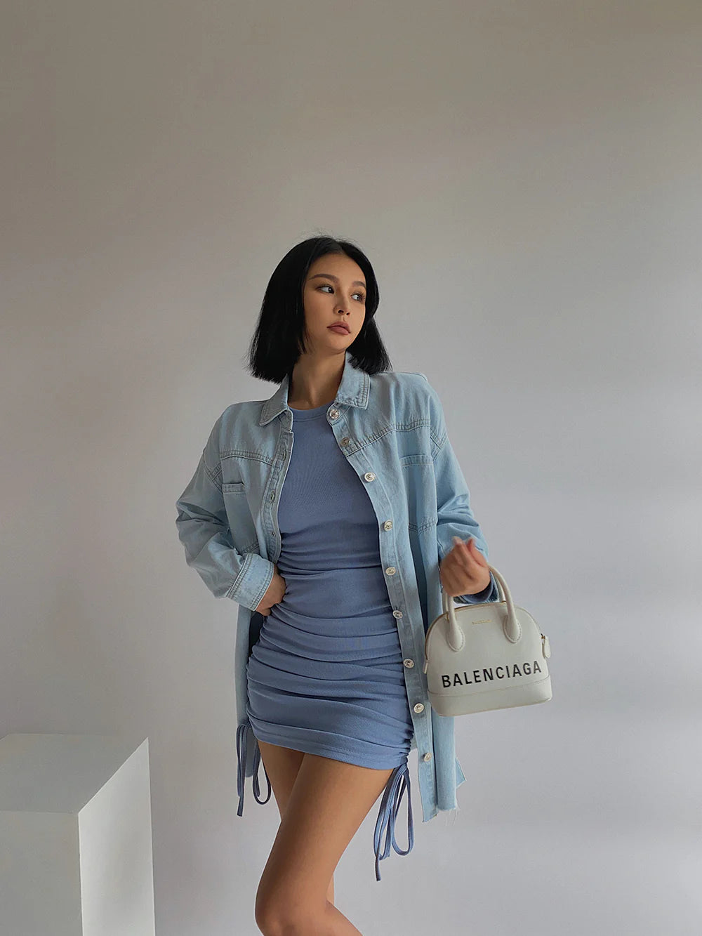Woman wearing a denim jacket over a blue dress, holding a Balenciaga handbag against a plain background.