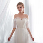 Woman wearing a white off-shoulder dress with sheer sleeves in front of white curtains.