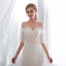 Woman wearing a white off-shoulder dress with sheer sleeves in front of white curtains.