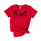Red t-shirt with 'Be still and know that I am God' text on a wooden surface with shoes and jeans