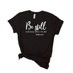 Black t-shirt with 'Be still and know that I am God' text, surrounded by jeans and sneakers on a white background