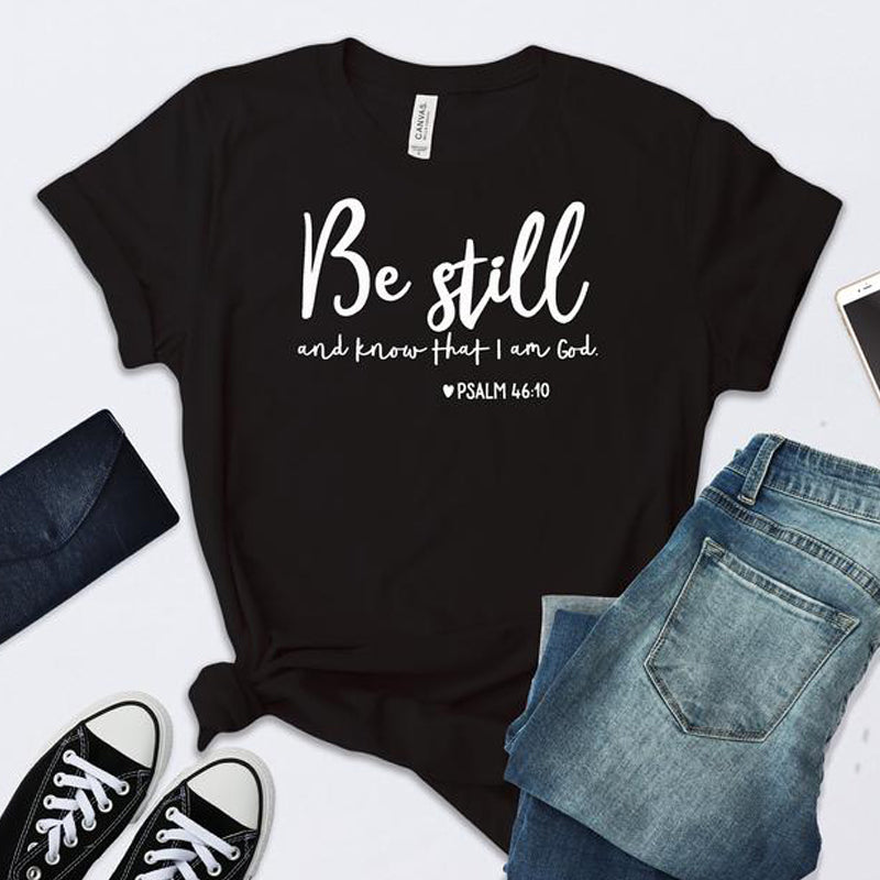 Black t-shirt with 'Be still and know that I am God' text, surrounded by jeans and sneakers on a white background