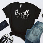 Black t-shirt with 'Be still and know that I am God' text, surrounded by jeans and sneakers on a white background